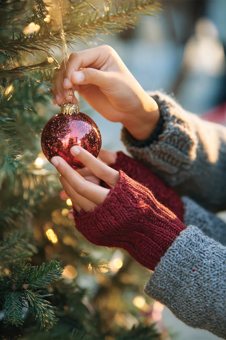 A child's hand and a parent's hand hanging a glass ornament on a lit holiday tree